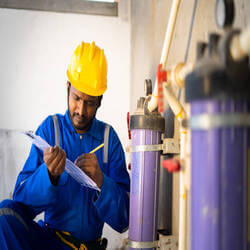a worker in a blue uniform and yellow hard hat inspecting some sort of machinery while holding a clipboard. 